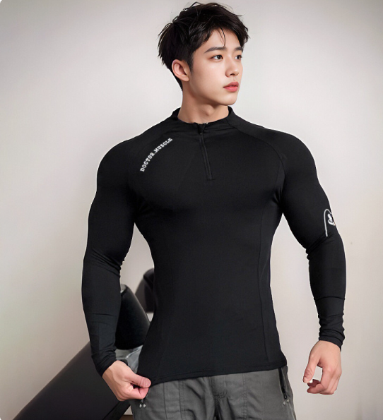 A muscular man wearing a tight-fitting black long-sleeve quarter-zip athletic shirt with white printed logos, paired with gray pants, posing against a neutral light gray background.